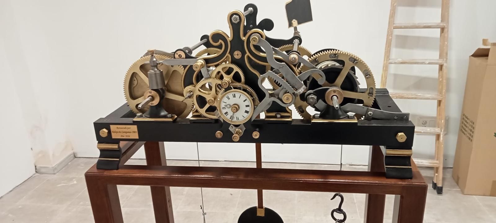 Engranaje mecánico antiguo sobre una mesa de madera en una habitación, con engranajes de latón y una esfera de reloj visible.
