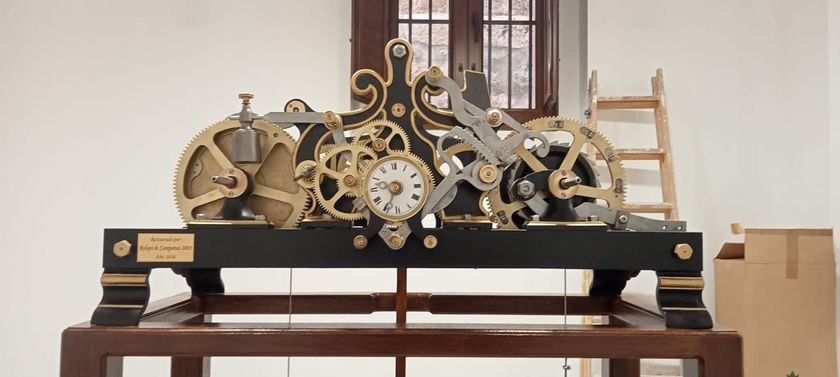 Reloj mecánico antiguo y ornamentado con engranajes a la vista sobre una mesa de exposición en un taller.