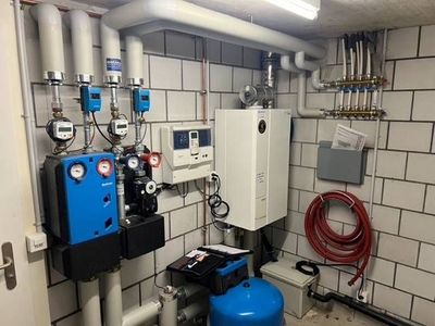 Industrieller Versorgungsraum mit Pumpen, Messgeräten, Schaltkästen und einem blauen Tank auf gefliestem Boden.