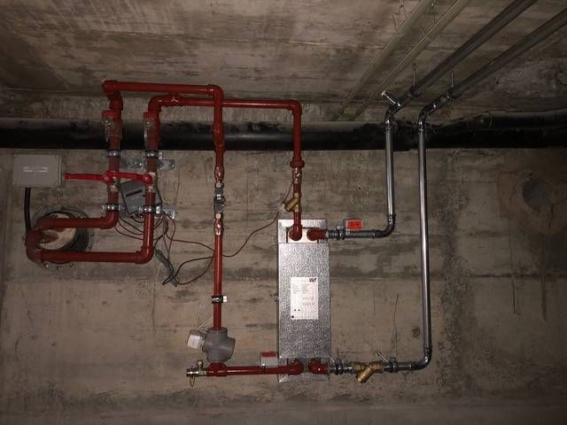 Kellerdecke mit roten und schwarzen Industrierohren und einer metallenen Steuereinheit an einer Betonwand