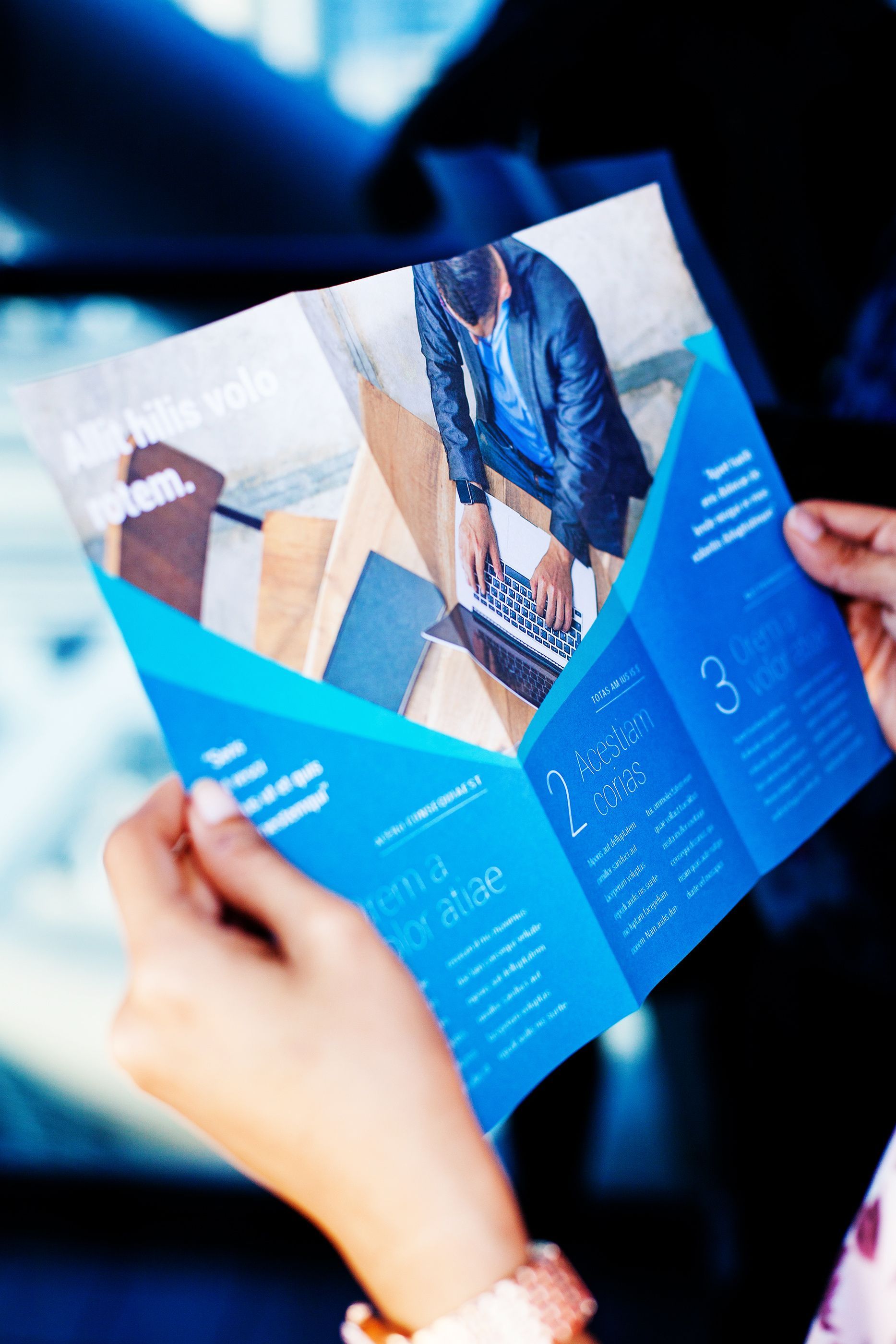 Mains tenant une brochure à trois volets ouverte ; design bleu, photo d'une personne travaillant sur un ordinateur portable.