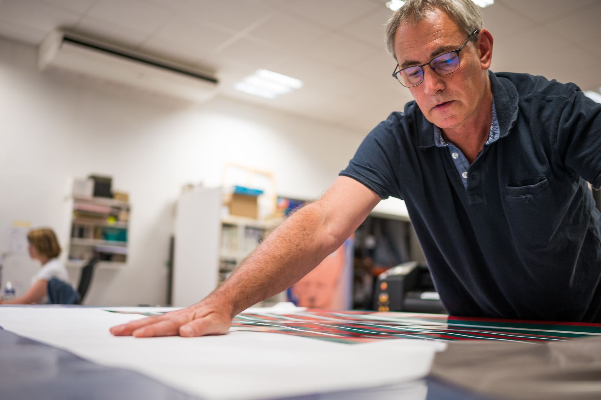 Un homme à lunettes, penché sur une table, examine une grande feuille de papier aux motifs colorés.