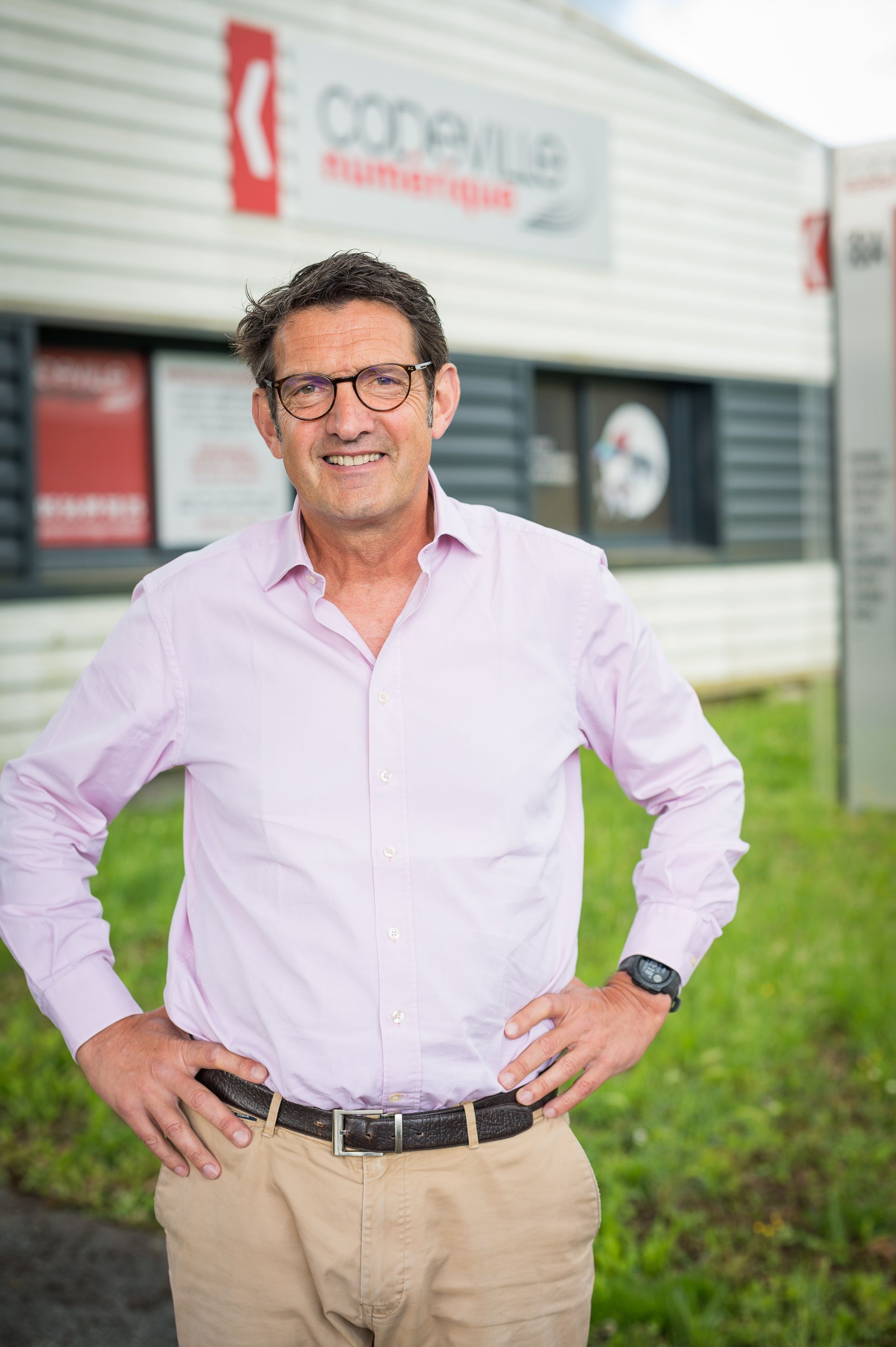 Un homme en chemise rose et lunettes, souriant, les mains sur les hanches, devant un bâtiment portant l'inscription « Codeville ».