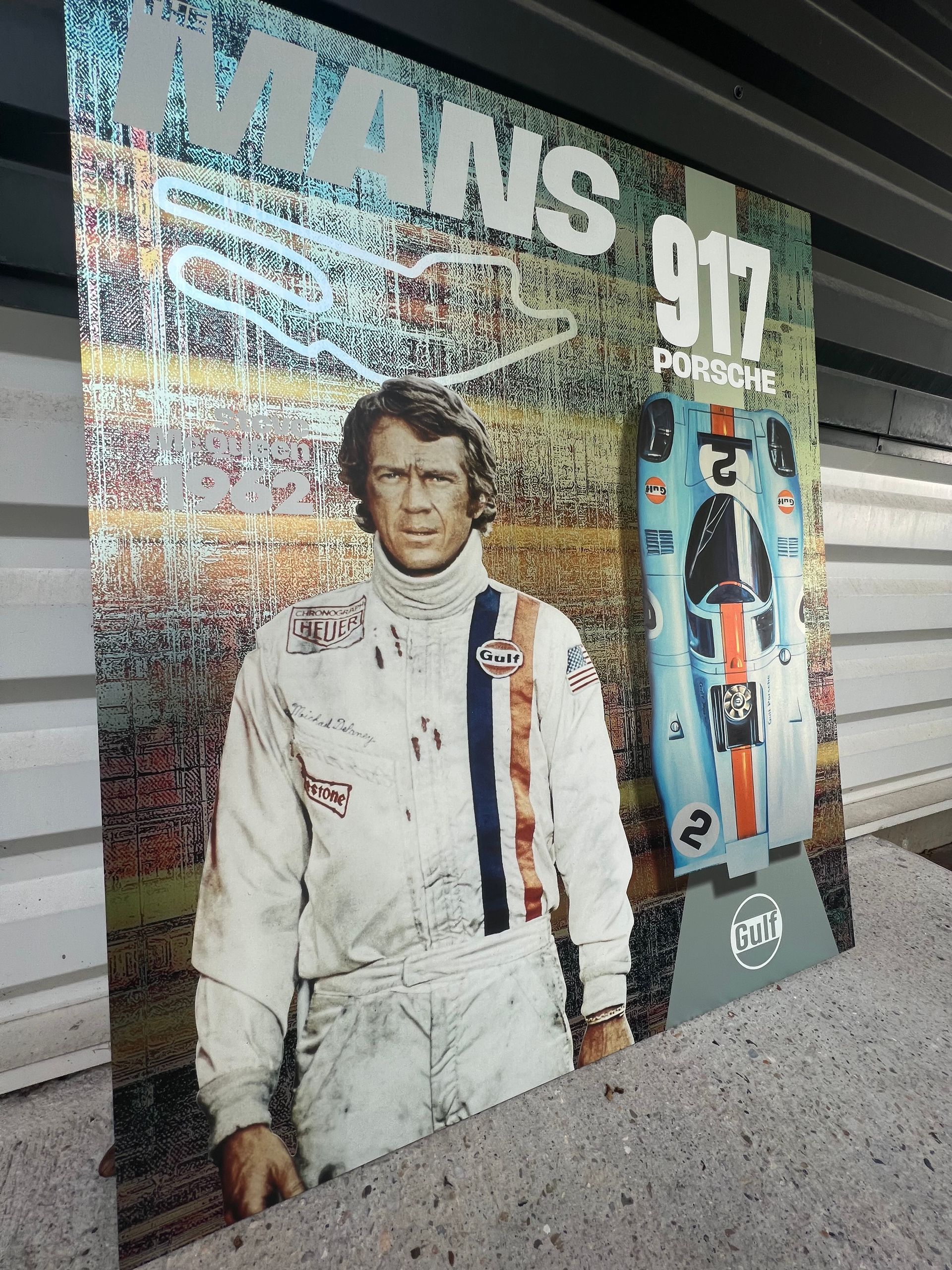 Steve McQueen, en combinaison de pilote, se tient à côté d'une Porsche 917 de course sur l'affiche du film « Le Mans ».