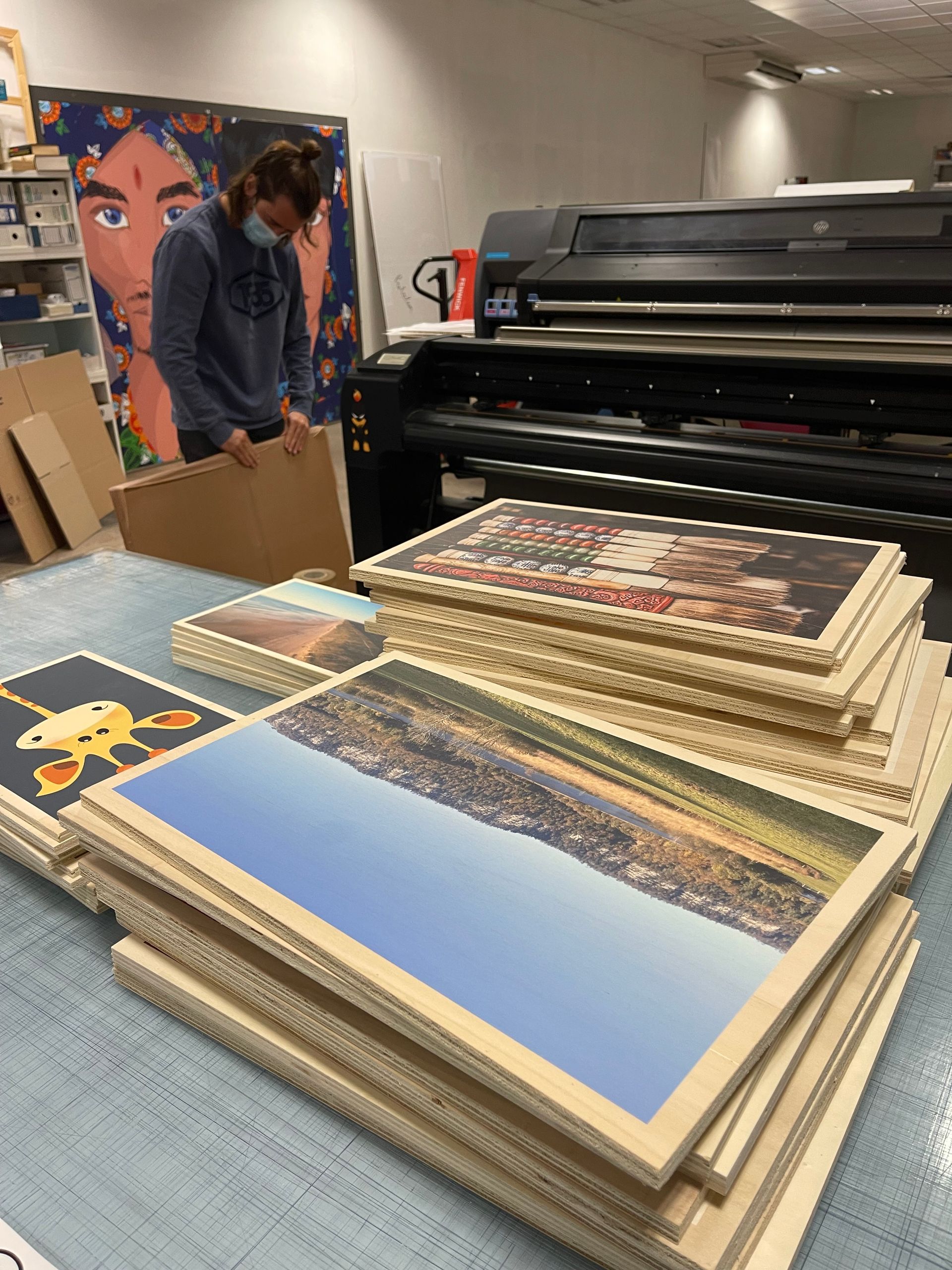 Une personne portant un masque prépare des tirages ; des piles d'images encadrées sur une table ; une imprimante grand format en arrière-plan.