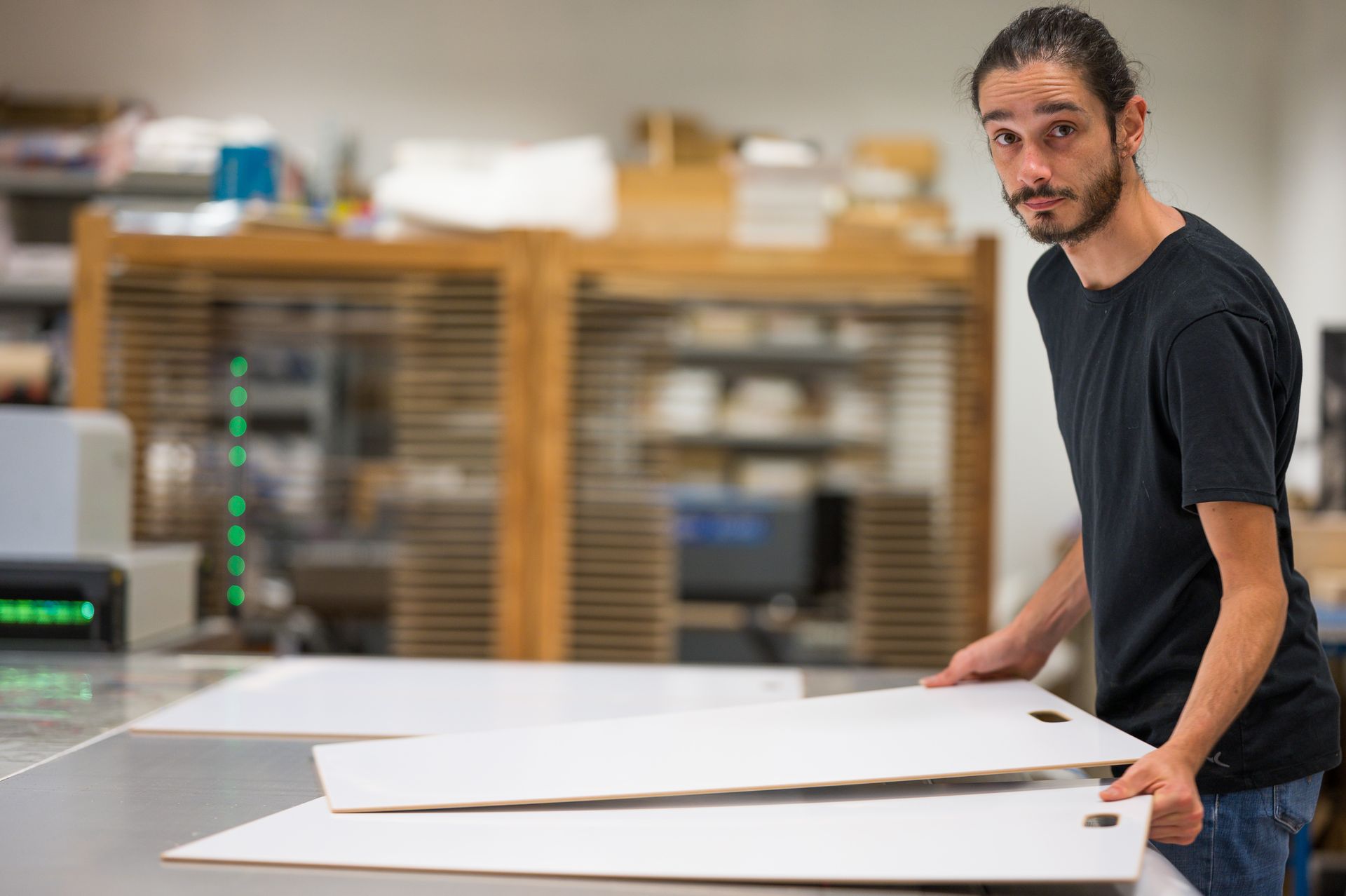 Un homme tenant des tableaux blancs regarde la caméra dans un atelier où l'on aperçoit en arrière-plan des imprimantes et des étagères.
