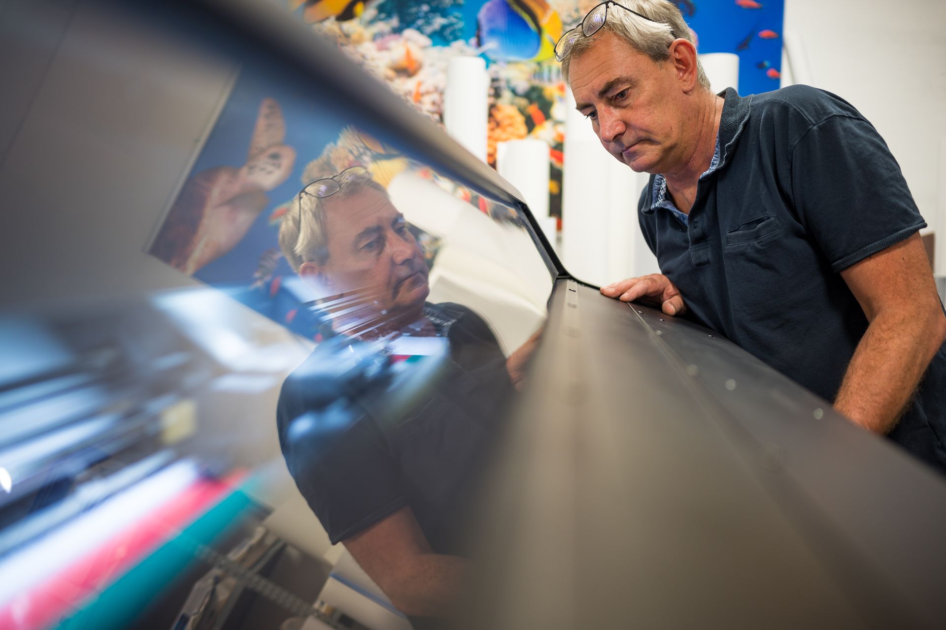 Un homme se reflète sur la surface d'une grande machine à imprimer qu'il est en train d'inspecter dans un décor d'atelier avec des œuvres d'art colorées visibles.