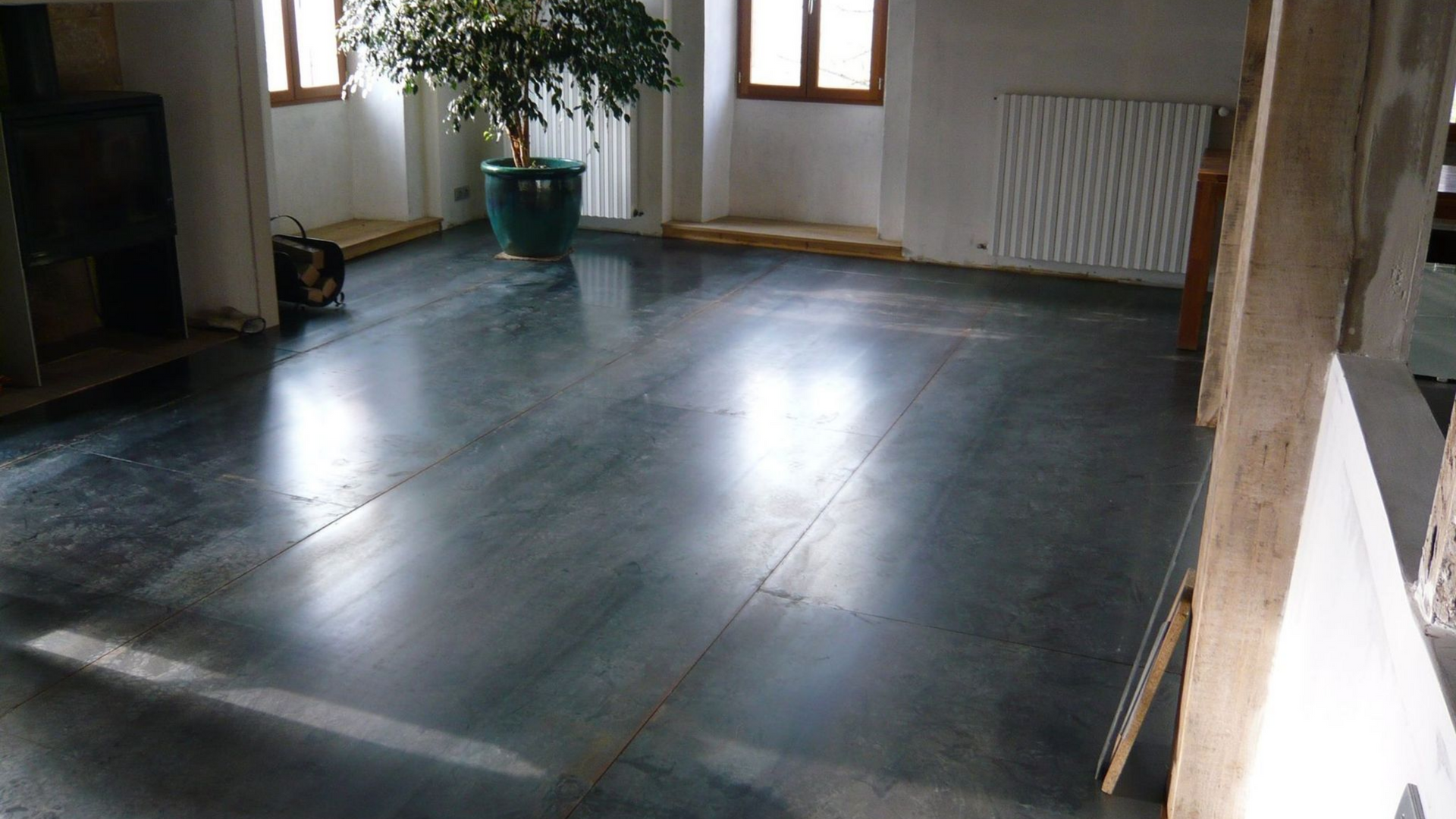Un salon avec un sol en métal gris brillant, un arbre en pot, des fenêtres et des poutres en bois apparentes.