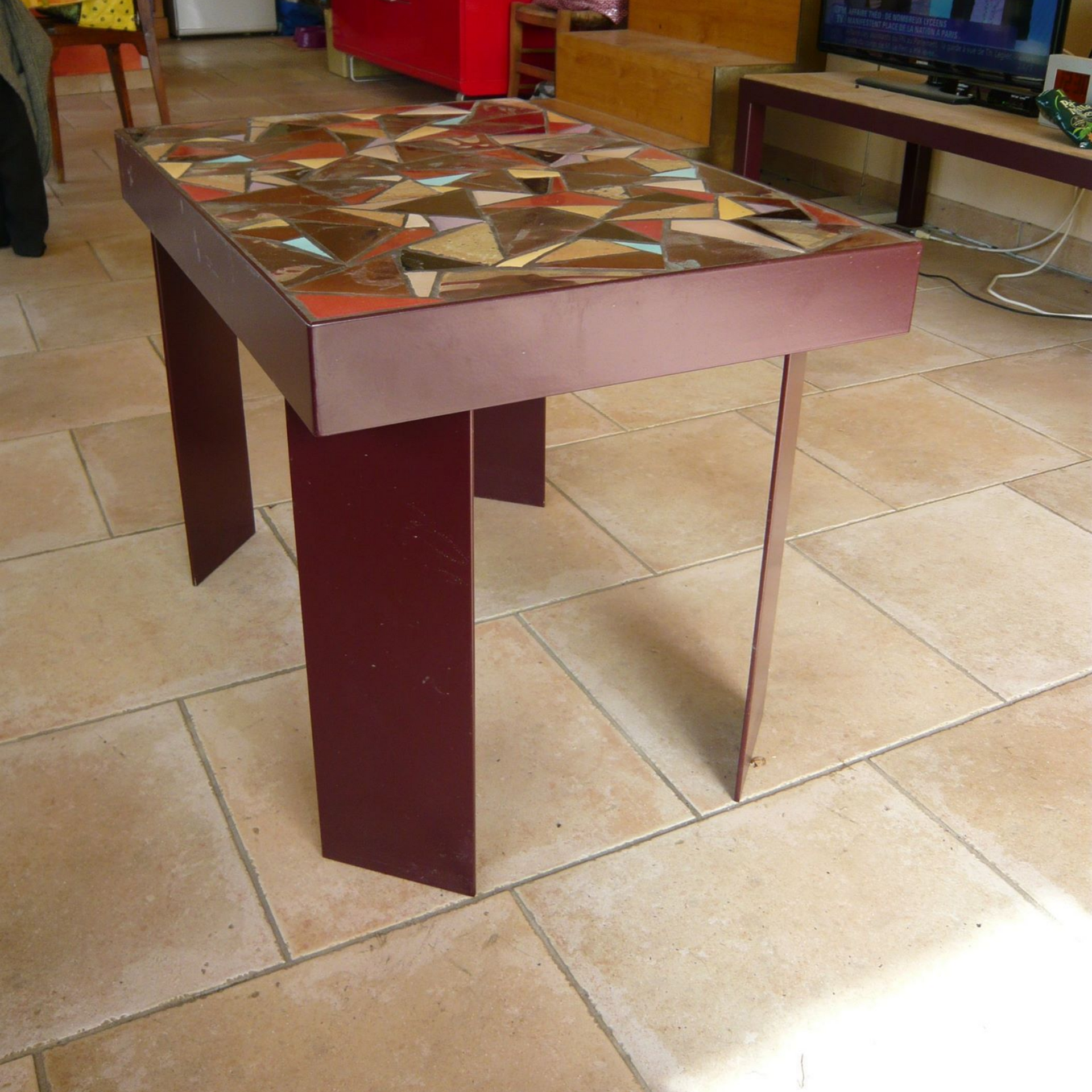 Table bordeaux à plateau en mosaïque, posée sur un sol carrelé.