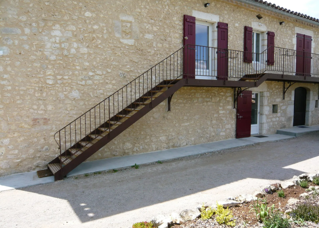 Bâtiment extérieur avec escalier métallique menant à un balcon, des volets rouges et une allée de gravier.