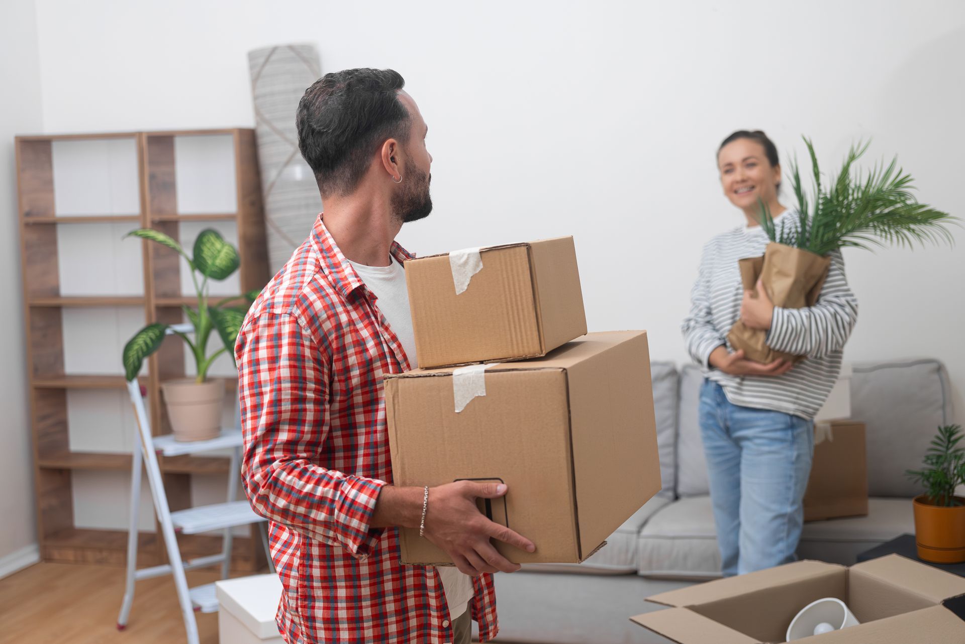 Deux personnes déplacent des cartons dans un salon lumineux, l'une d'elles tenant une plante et souriant.