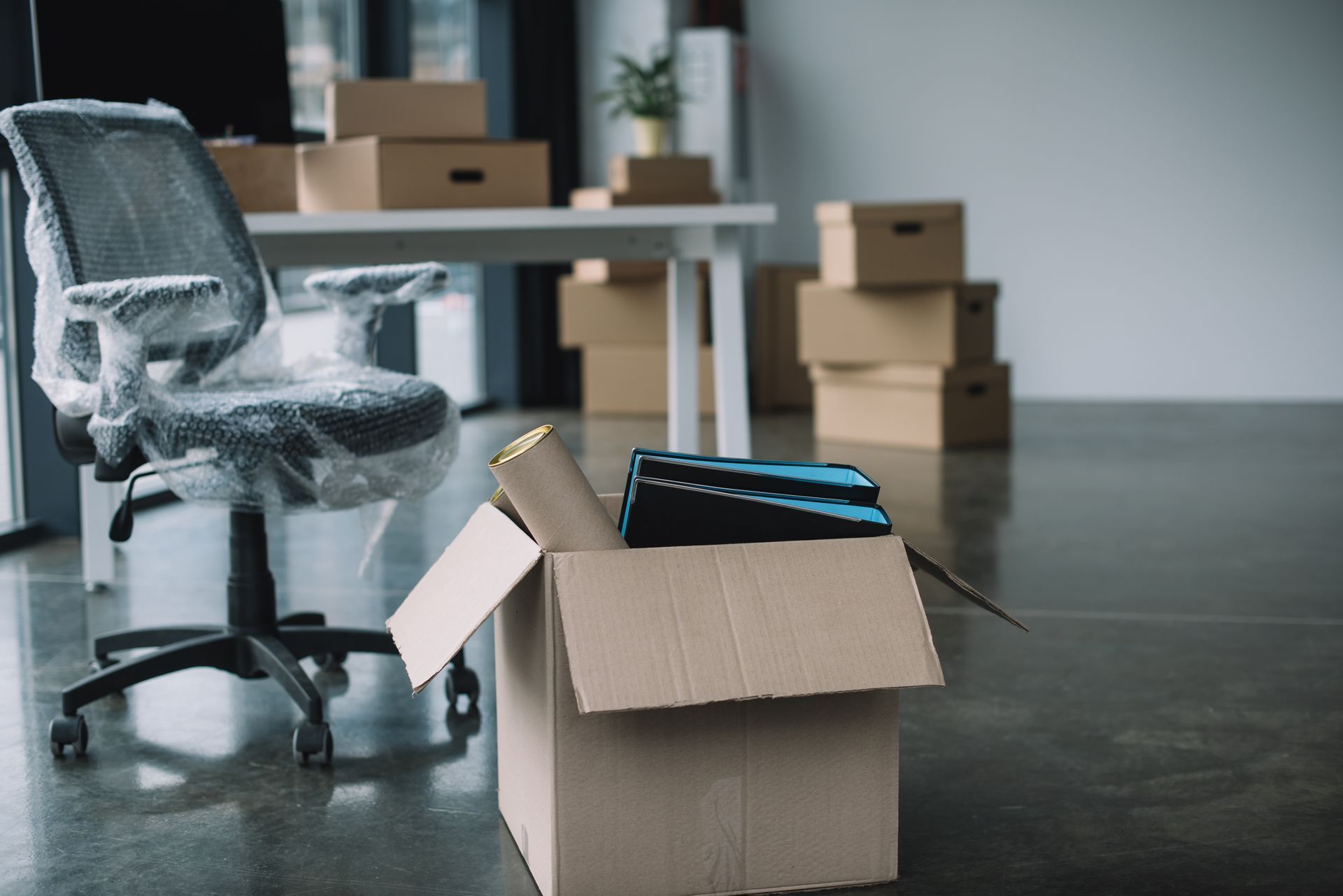 Dans une pièce dépouillée, une chaise de bureau emballée côtoie des cartons ouverts, suggérant un déménagement ou un entreposage.