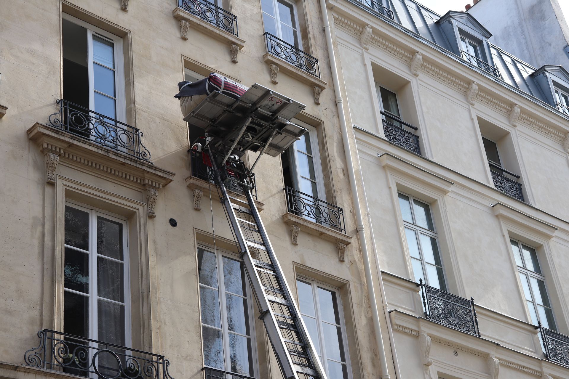 Des pompiers à bord d'un camion-échelle atteignent le balcon du dernier étage d'un immeuble d'appartements beige.