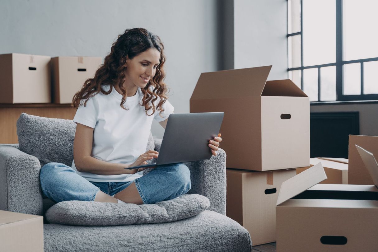 Une femme assise sur un canapé utilise un ordinateur portable au milieu de cartons de déménagement dans un salon lumineux.