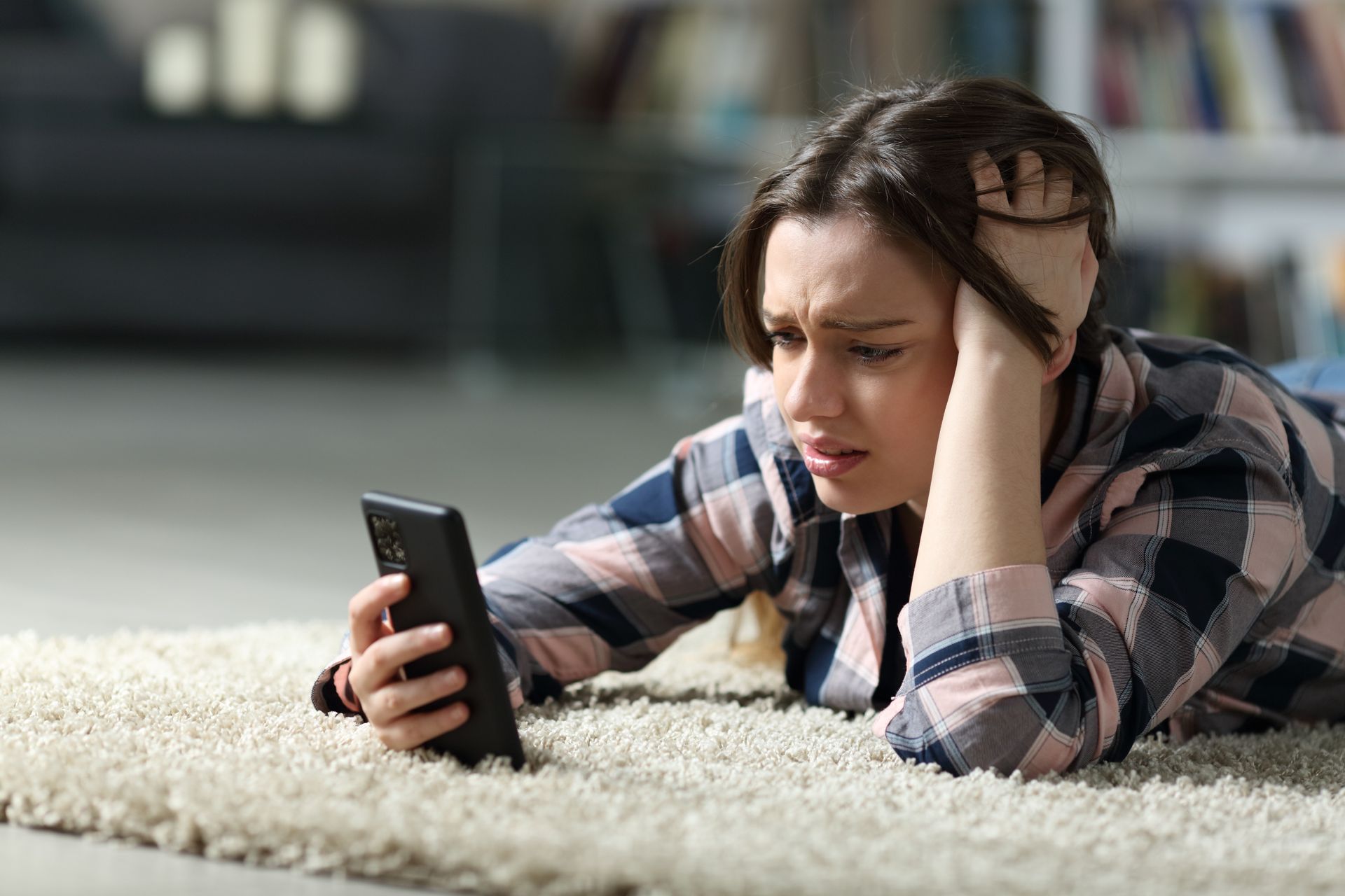 Une femme allongée sur un tapis, regardant son téléphone avec une expression inquiète, la main sur la tête.
