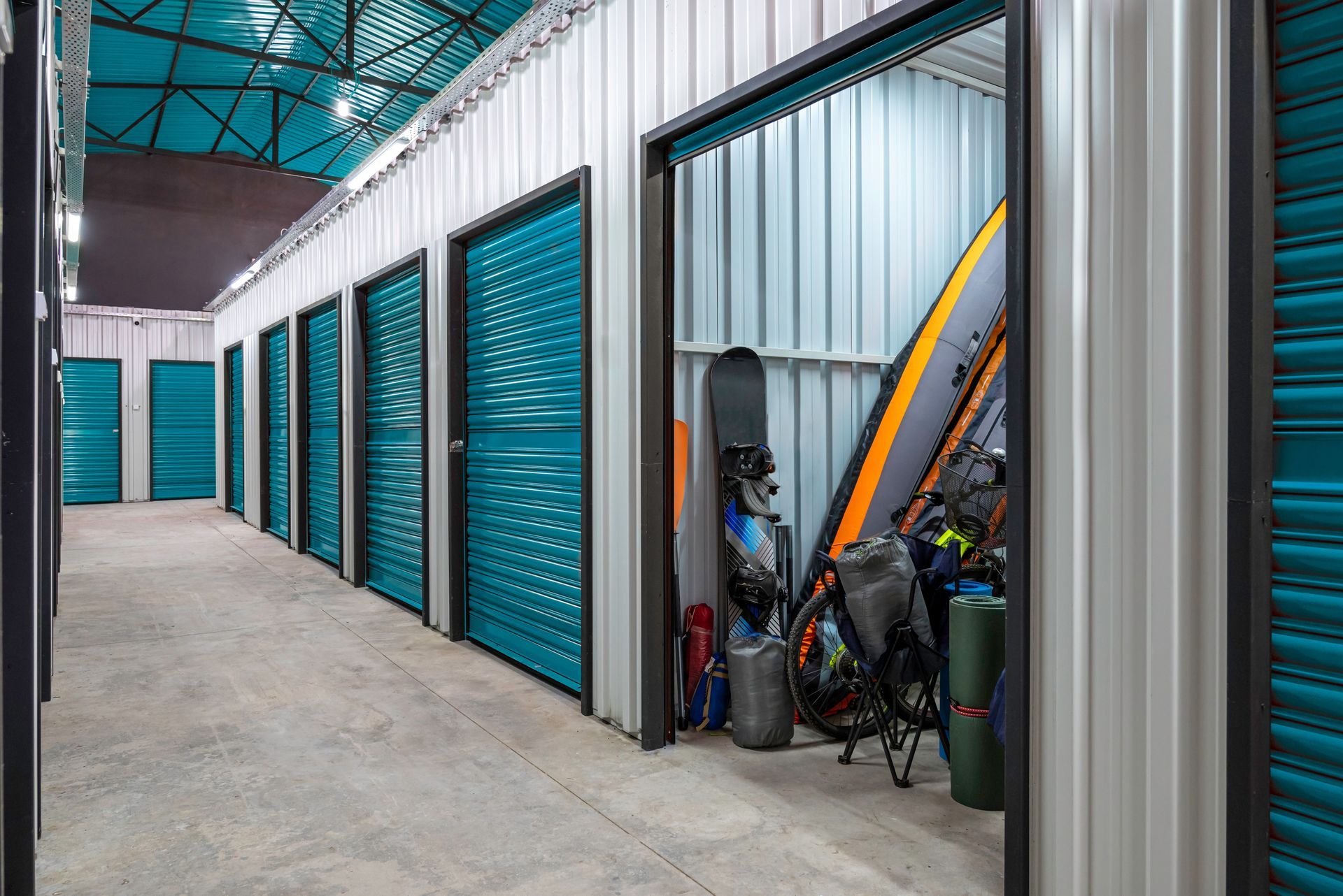 Couloir d'un box de stockage avec des portes turquoise et quelques vélos, avec une planche de surf appuyée contre un box ouvert.