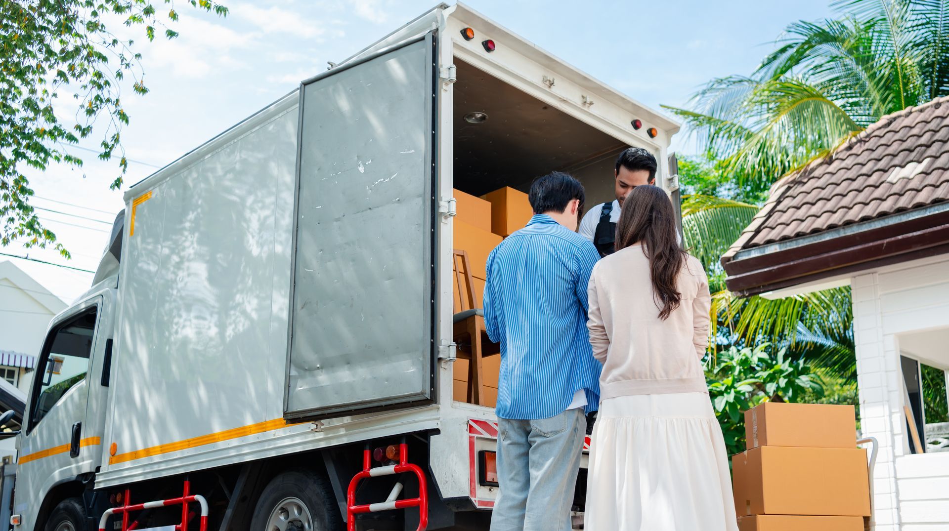 Un camion de livraison décharge des cartons à côté d'une maison, deux personnes se tenant à la porte de chargement ouverte.