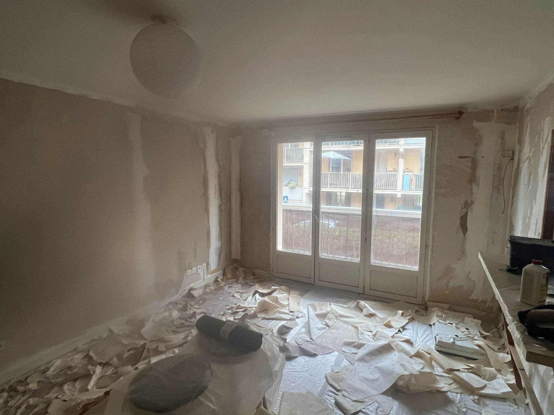 Chambre partiellement rénovée avec un sol jonché de débris et de grandes fenêtres laissant entrer la lumière du jour
