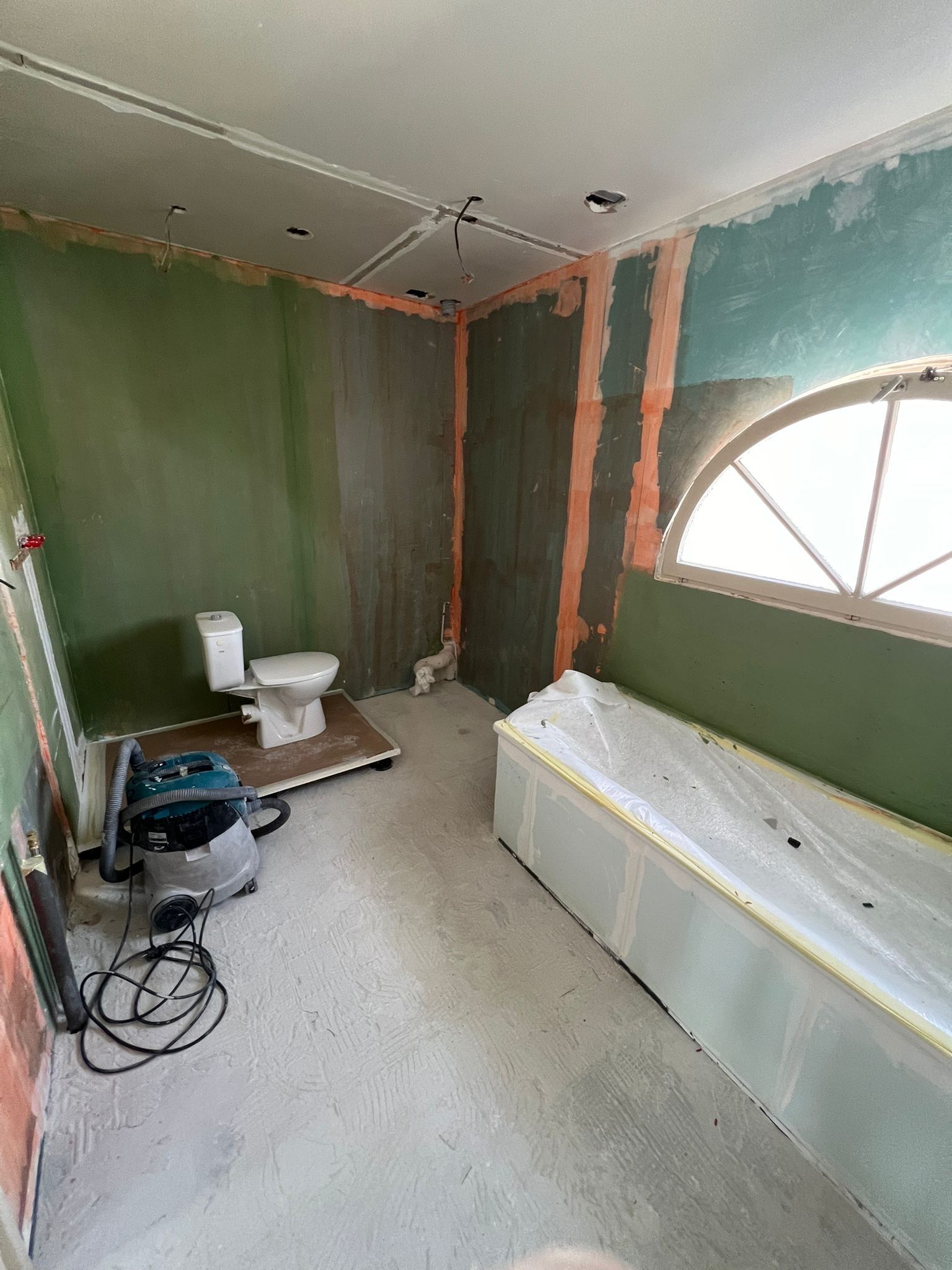 Salle de bains partiellement rénovée avec murs verts, toilettes et longue baignoire blanche sous une fenêtre en demi-cercle