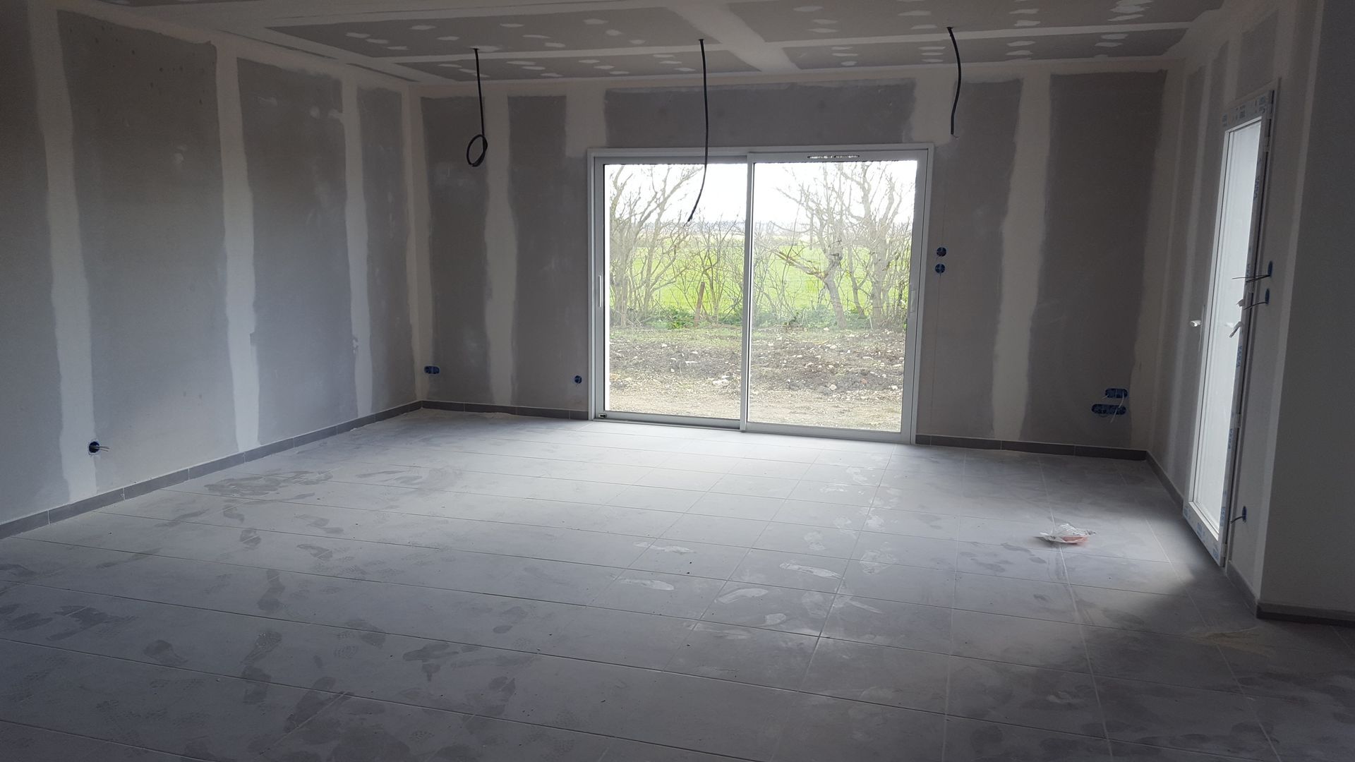 Pièce vide et inachevée, aux murs gris, cloisons sèches apparentes et portes coulissantes en verre donnant sur une cour verdoyante.
