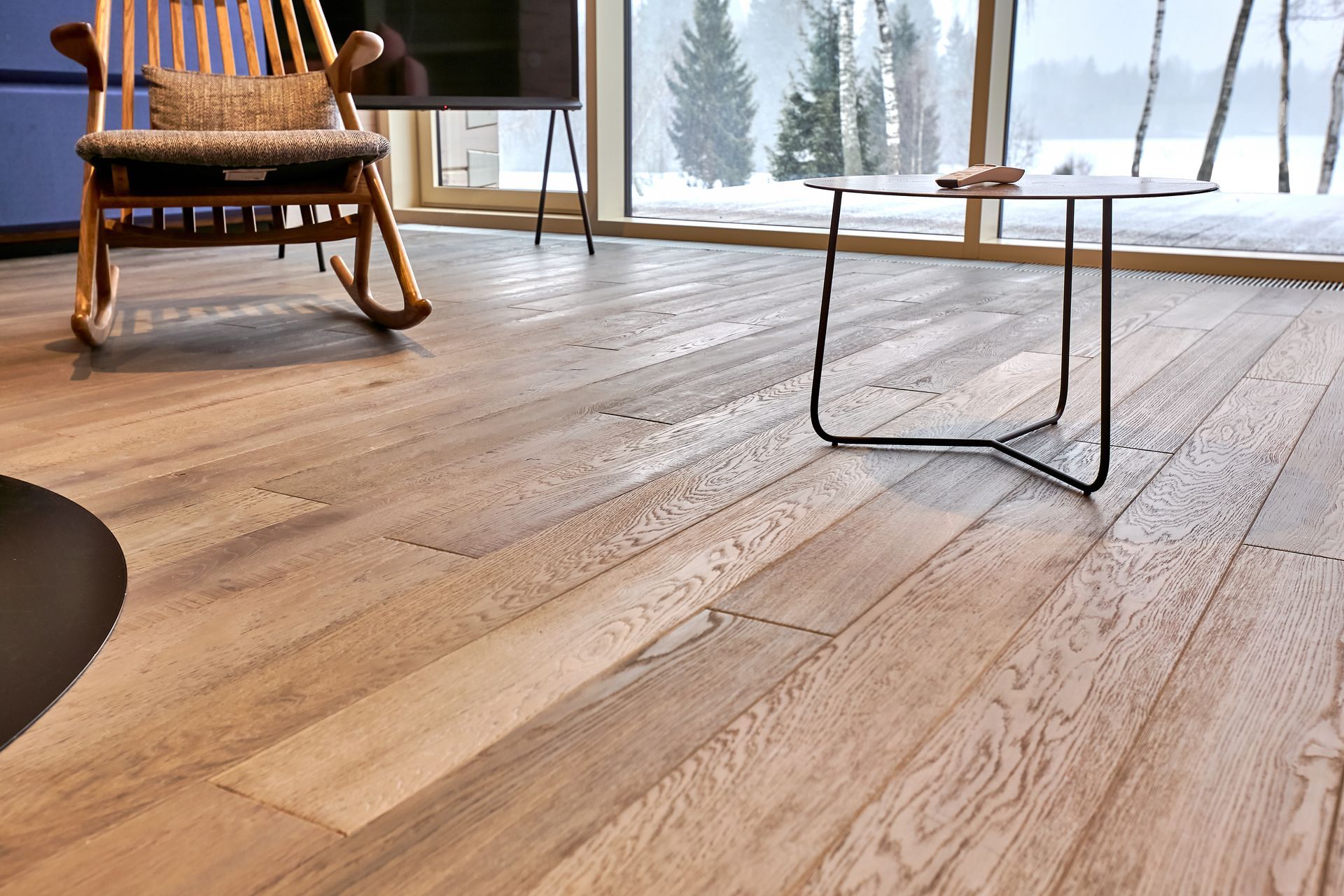 Salon moderne avec un fauteuil à bascule en bois, une table basse en verre et de grandes fenêtres donnant sur un paysage enneigé