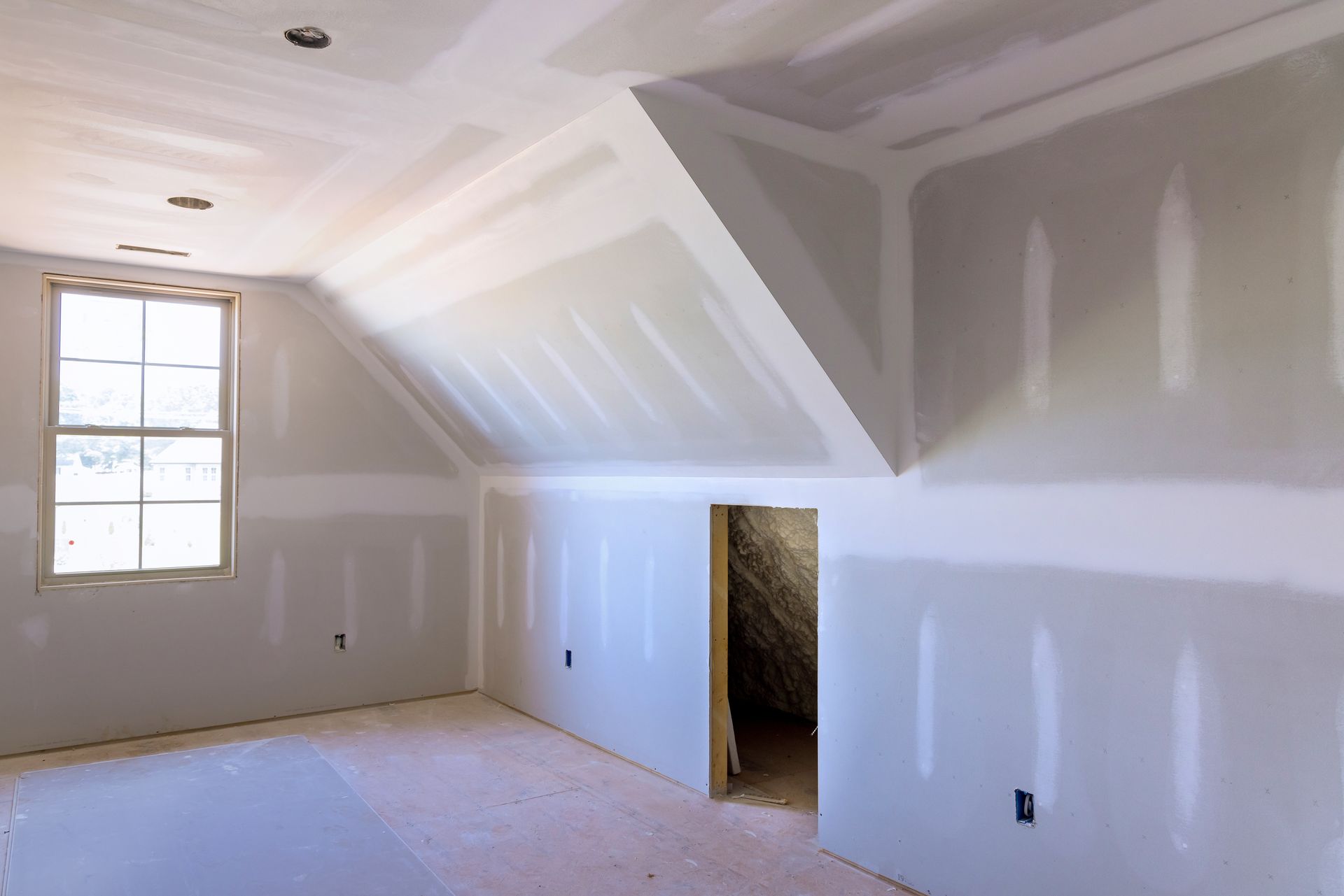 Pièce vide et inachevée avec cloisons blanches, plafond mansardé, fenêtre et porte ouverte donnant sur un escalier.