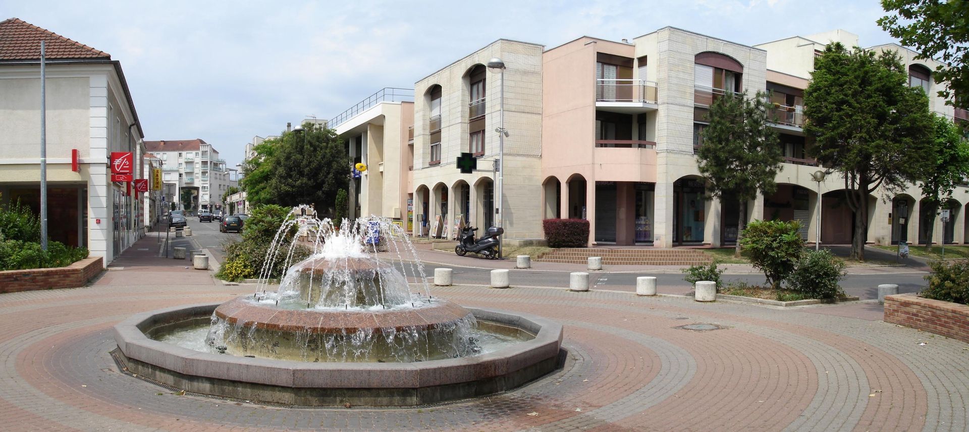 Place avec une fontaine circulaire et les bâtiments environnants par temps nuageux