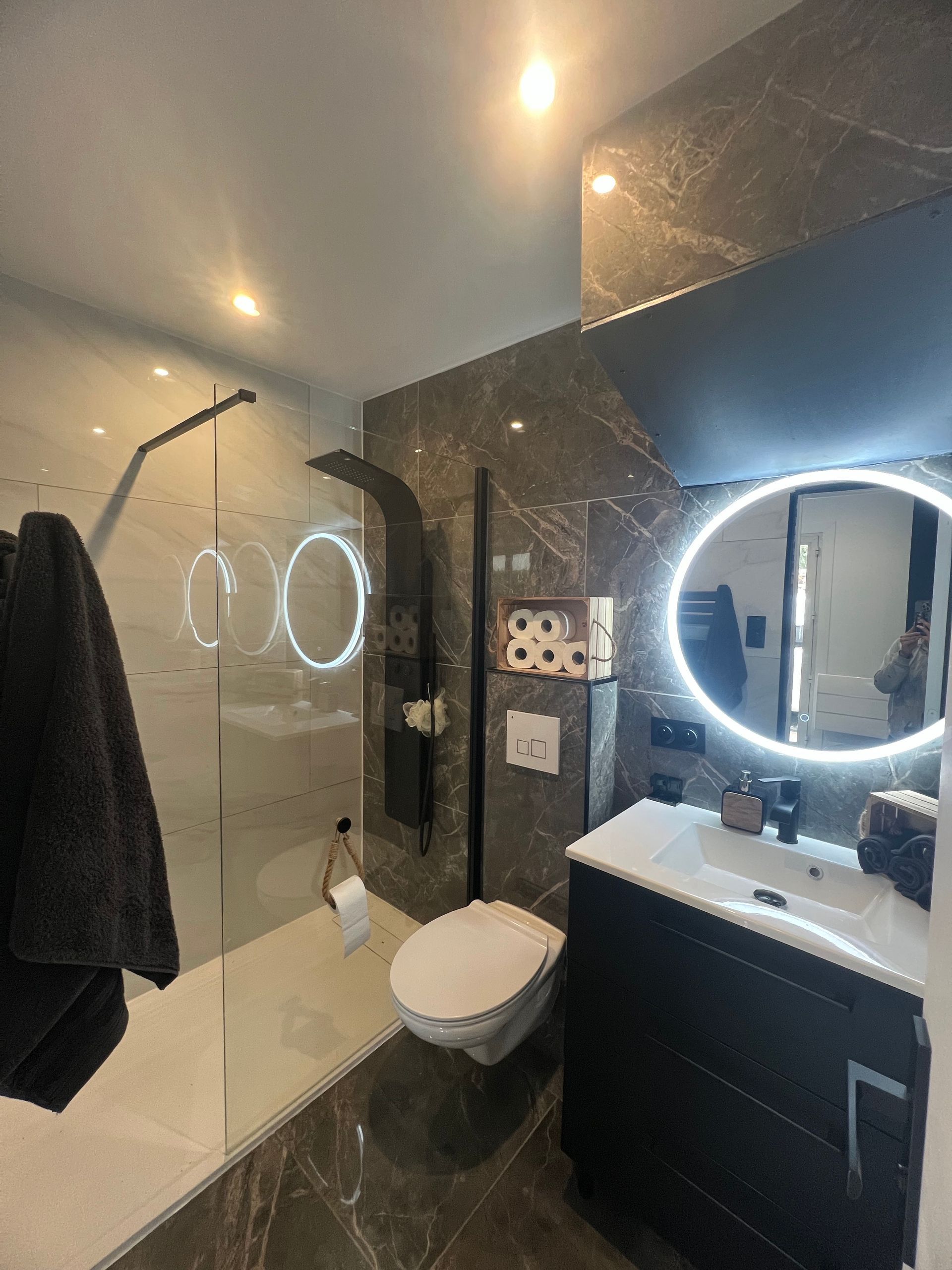 Salle de bains moderne avec douche à l'italienne, toilettes, carrelage foncé et miroir rond éclairé au-dessus d'un lavabo.