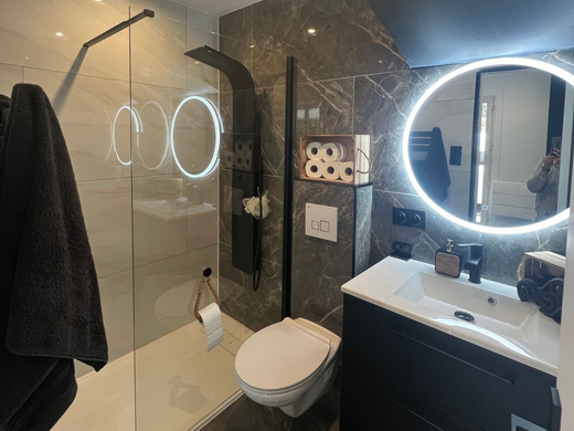 Salle de bains moderne avec miroir rond éclairé, meuble-lavabo noir, toilettes et cabine de douche en verre.