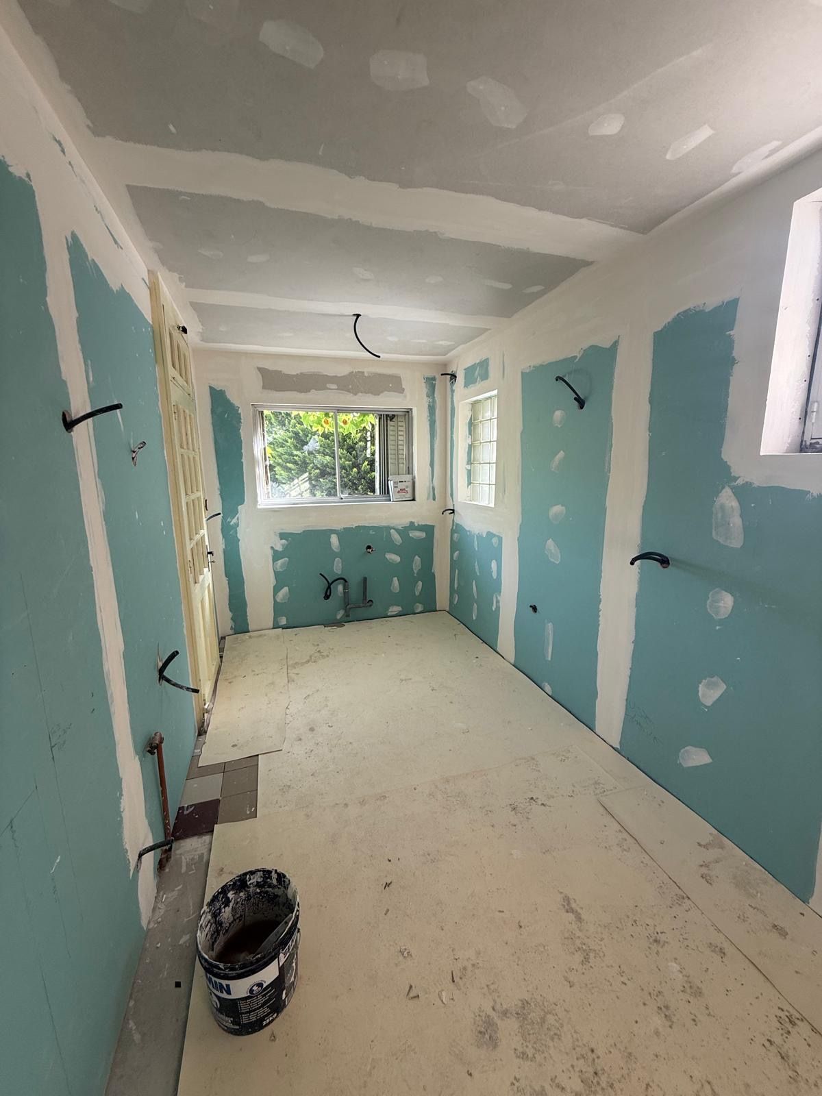 Pièce étroite en cours de rénovation avec des panneaux de plaques de plâtre bleus, des zones blanches au plafond et une fenêtre au fond.