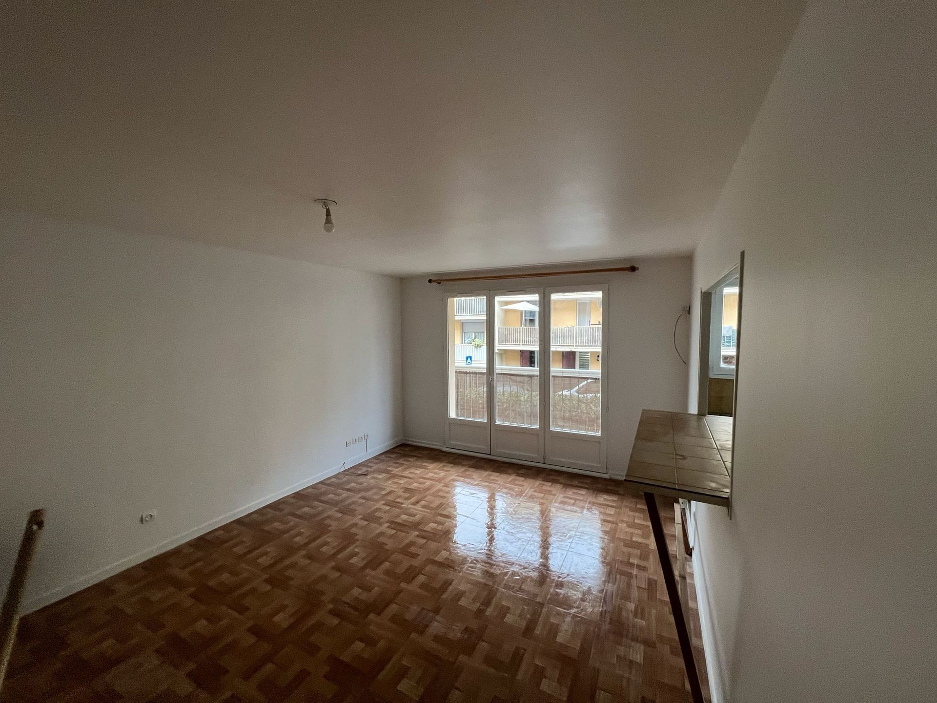 Salon vide avec parquet, murs blancs et grandes fenêtres au fond.