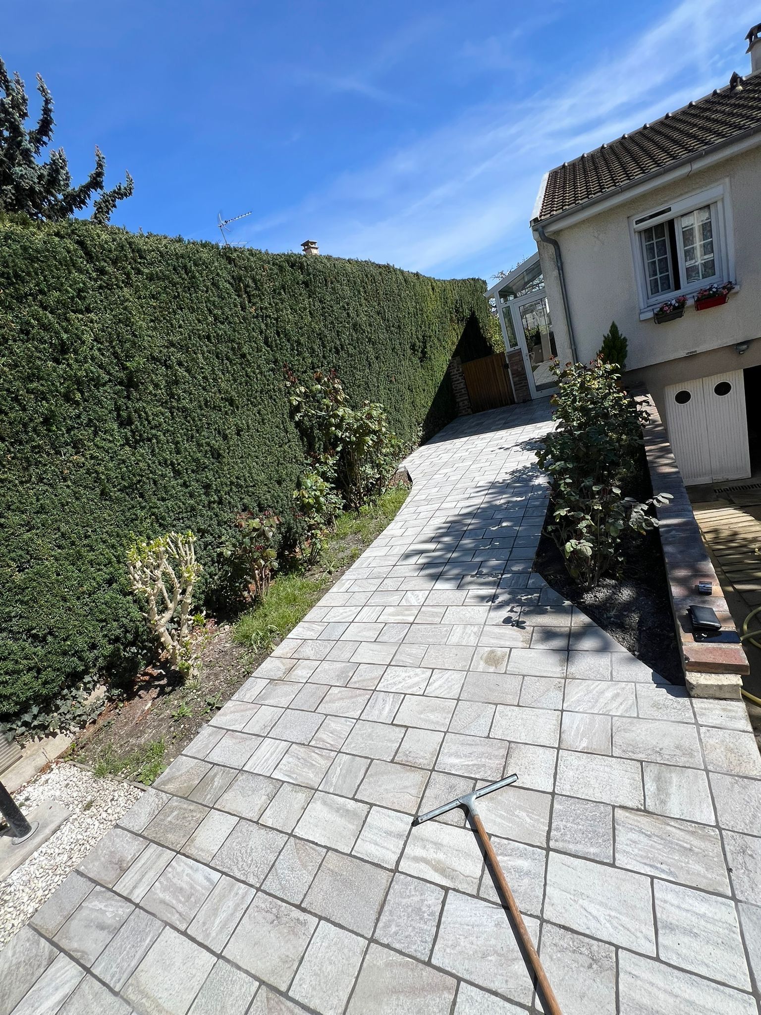 Allée pavée ensoleillée longeant une haute haie et une maison, avec un balai au premier plan.