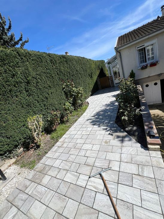Allée de jardin en pierre longeant de hautes haies et une maison sous un ciel bleu clair