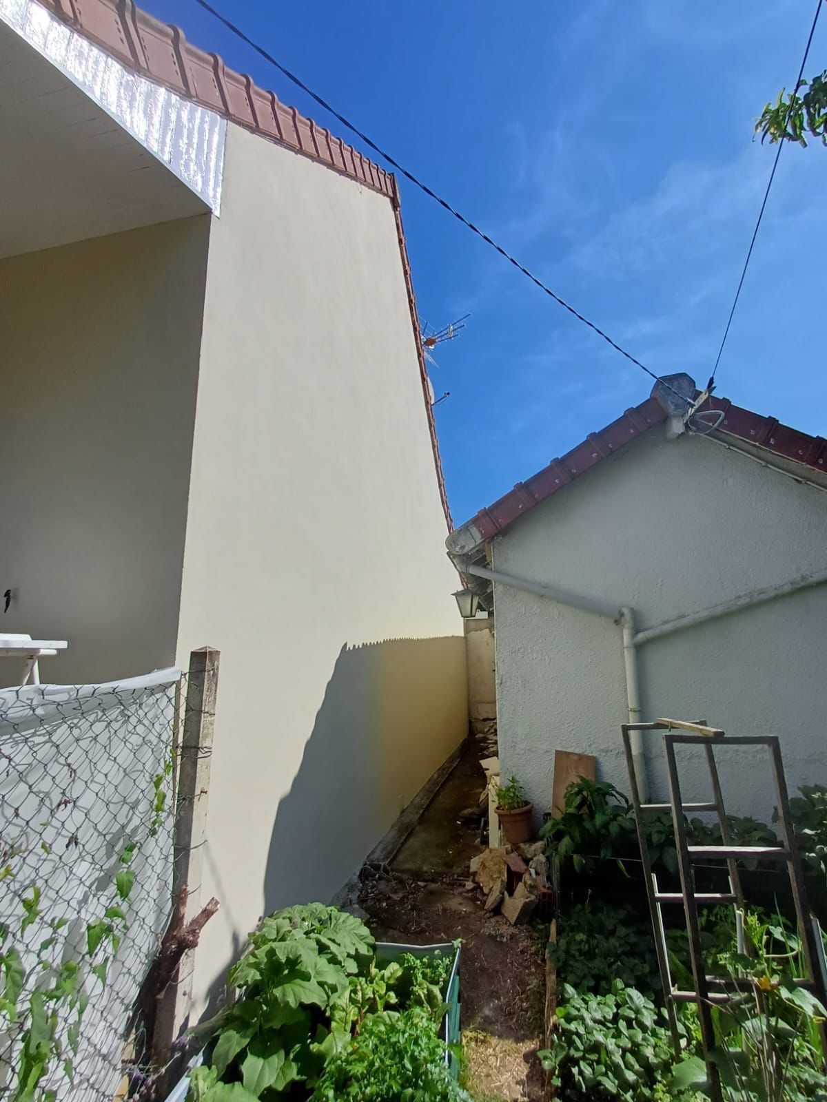 Étroit passage extérieur jouxtant une maison couleur crème et un petit jardin sous un ciel d'un bleu éclatant.