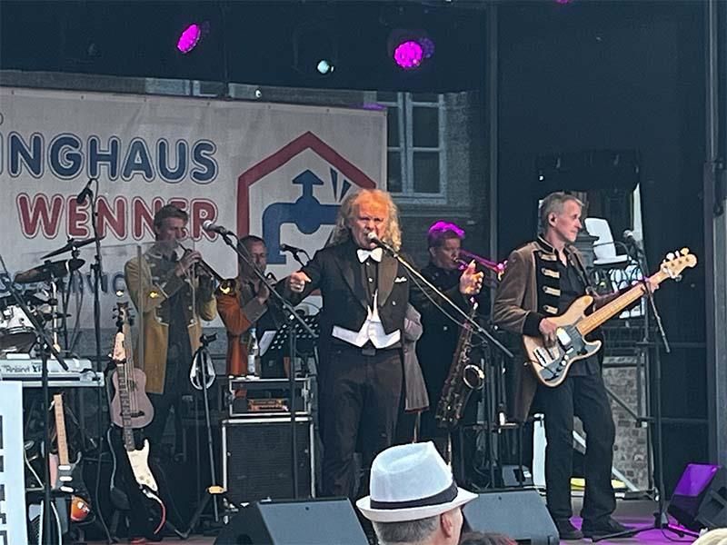 Eine Gruppe von Männern spielt Instrumente auf einer Bühne vor einem Schild mit der Aufschrift „Inghaus Wenner“