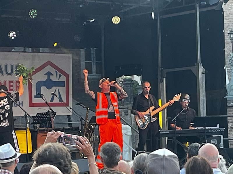 Ein Mann in einer orangefarbenen Weste singt vor einer Menschenmenge in ein Mikrofon.