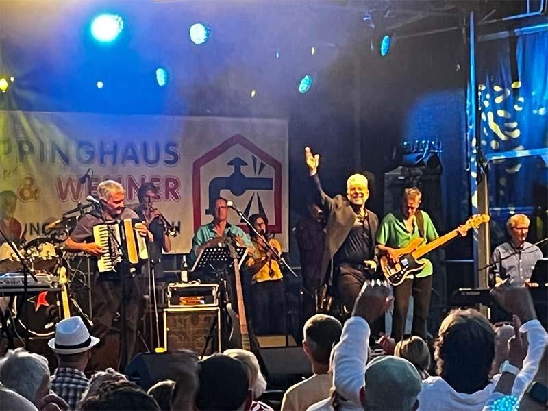 Eine Gruppe von Männern spielt Instrumente auf einer Bühne vor einem Schild mit der Aufschrift „Pinghaus“