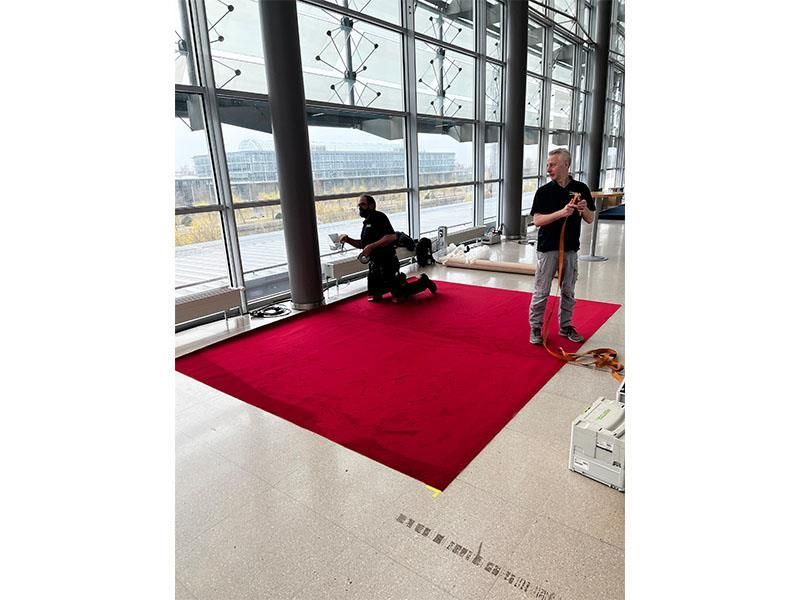 Zwei Männer knien auf einem roten Teppich vor einem großen Fenster.