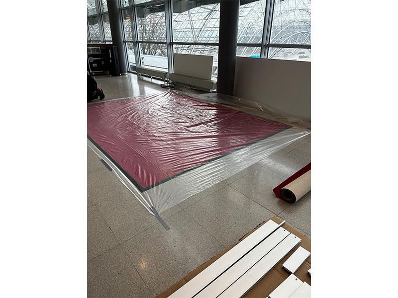 In einem Raum mit vielen Fenstern wird ein roter Teppich ausgerollt.