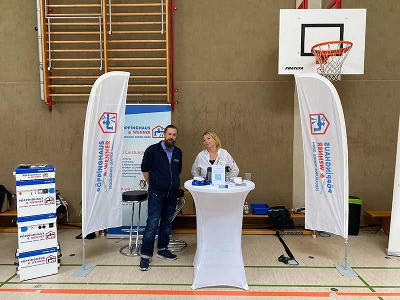 Ein Mann und eine Frau stehen hinter einem Tisch in einer Turnhalle mit einem Basketballkorb im Hintergrund
