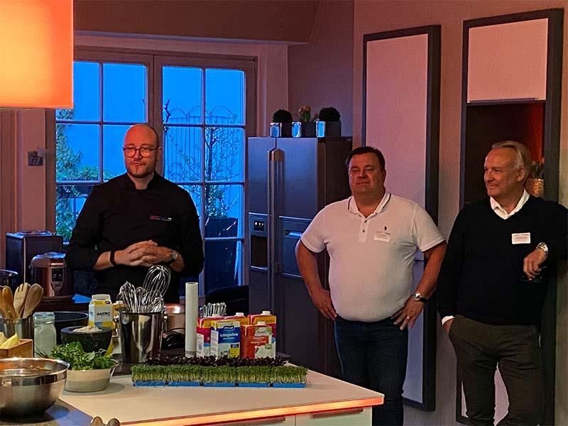 Drei Männer stehen in einer Küche und schauen etwas an