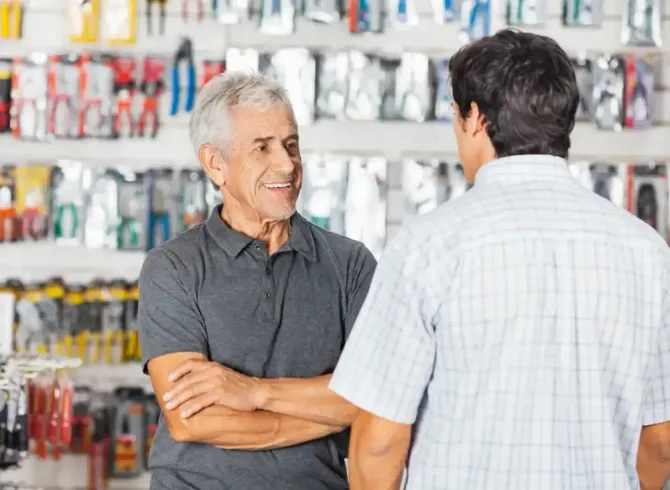 Empleado de tienda hablando con un cliente en una tienda; productos en estantes en el fondo.