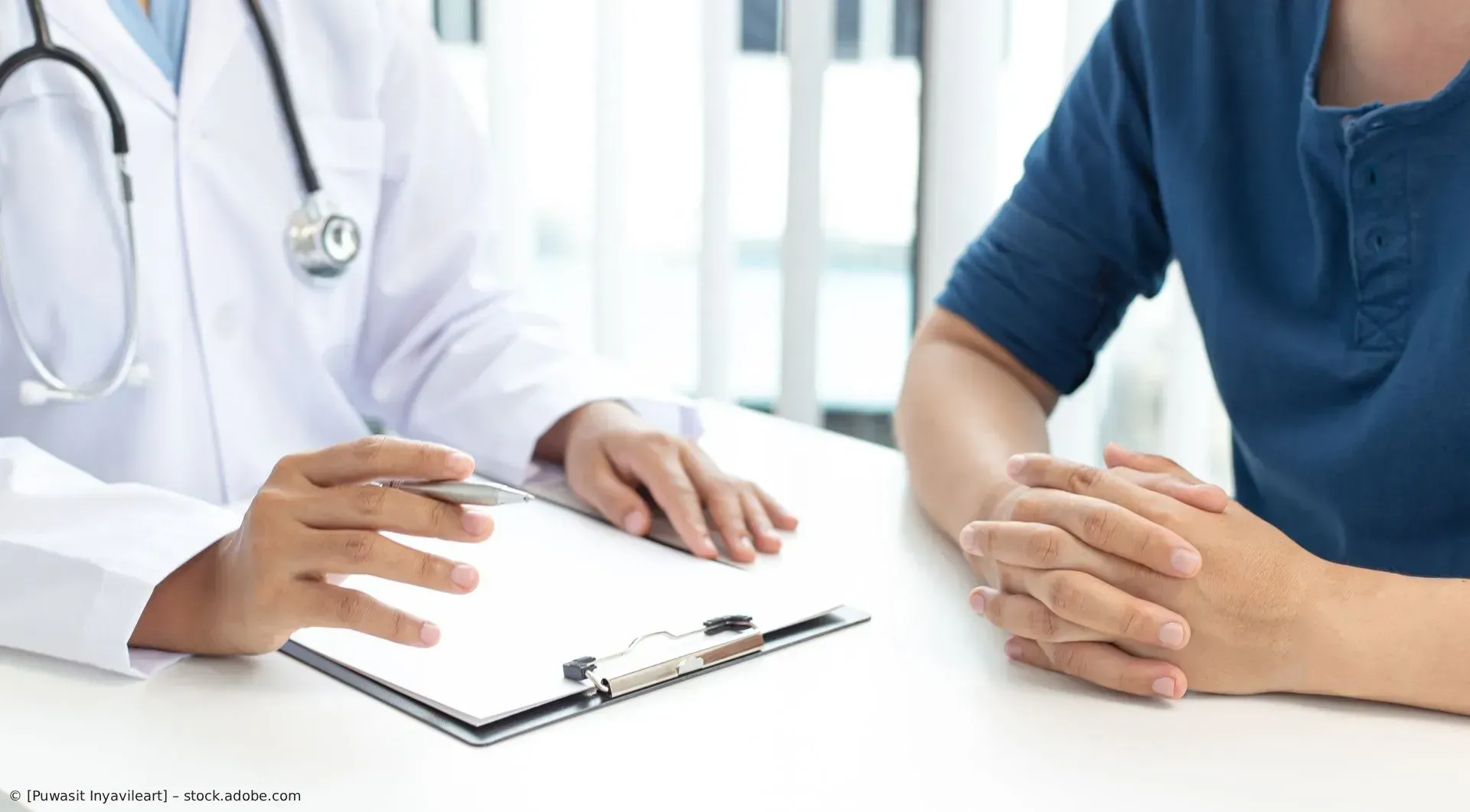 Ein Arzt unterhält sich mit einem Patienten an einem Schreibtisch, die Hände ineinander verschränkt,