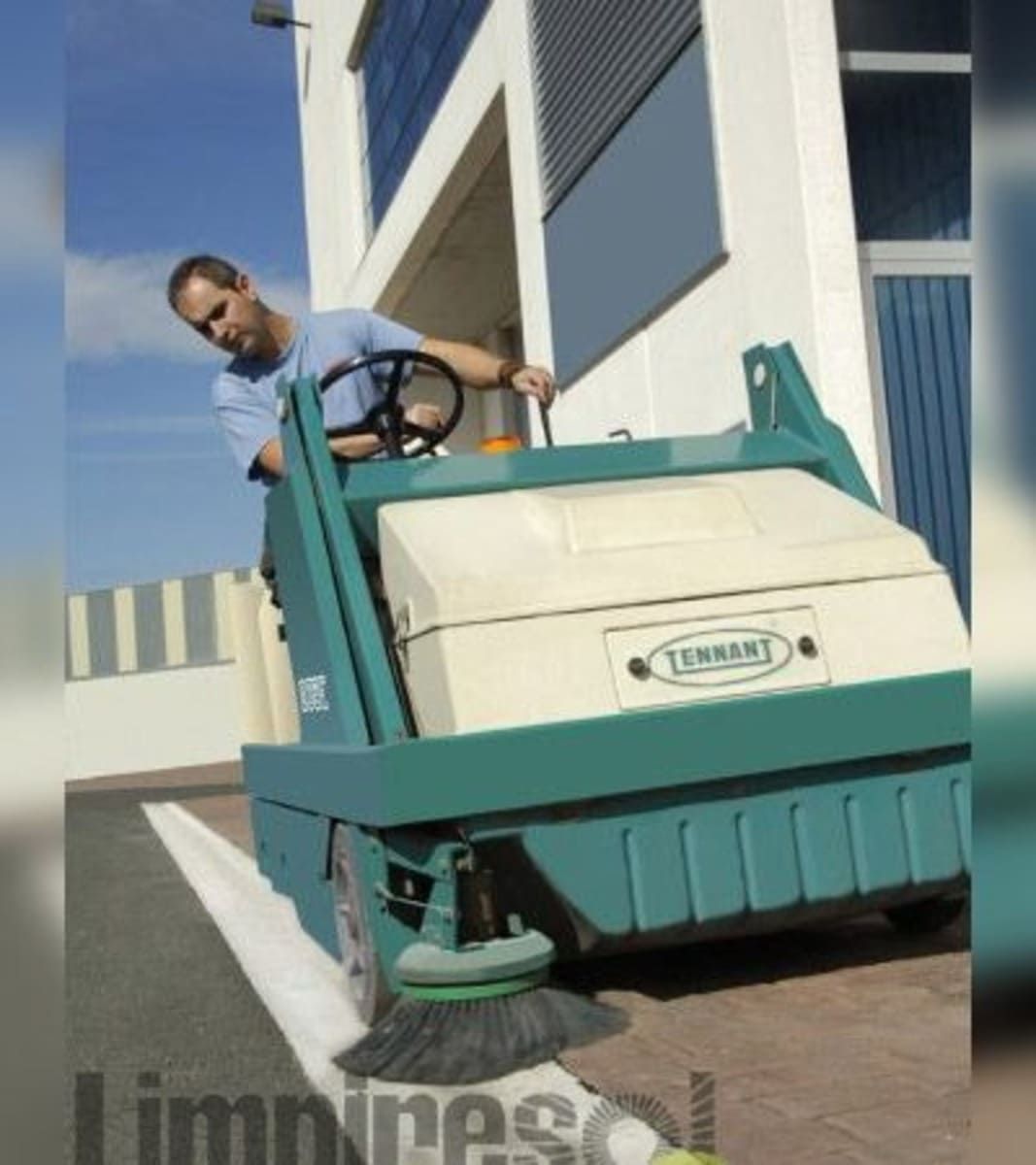 Un trabajador en una barredora verde limpia una zona pavimentada junto a un edificio.