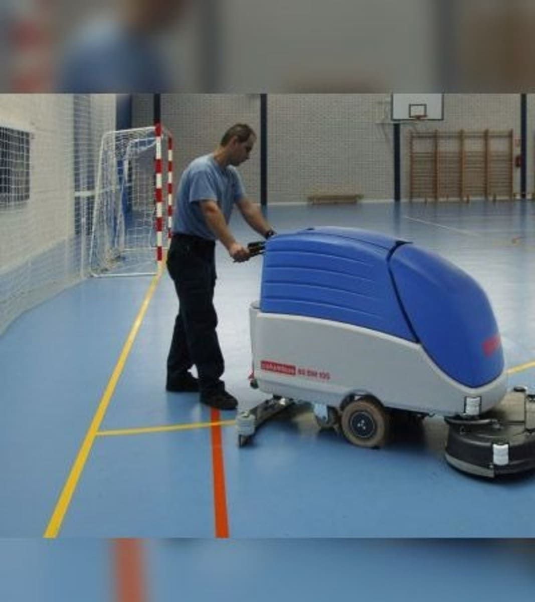 Un hombre empuja una máquina azul de limpieza de suelos en un gimnasio cubierto.