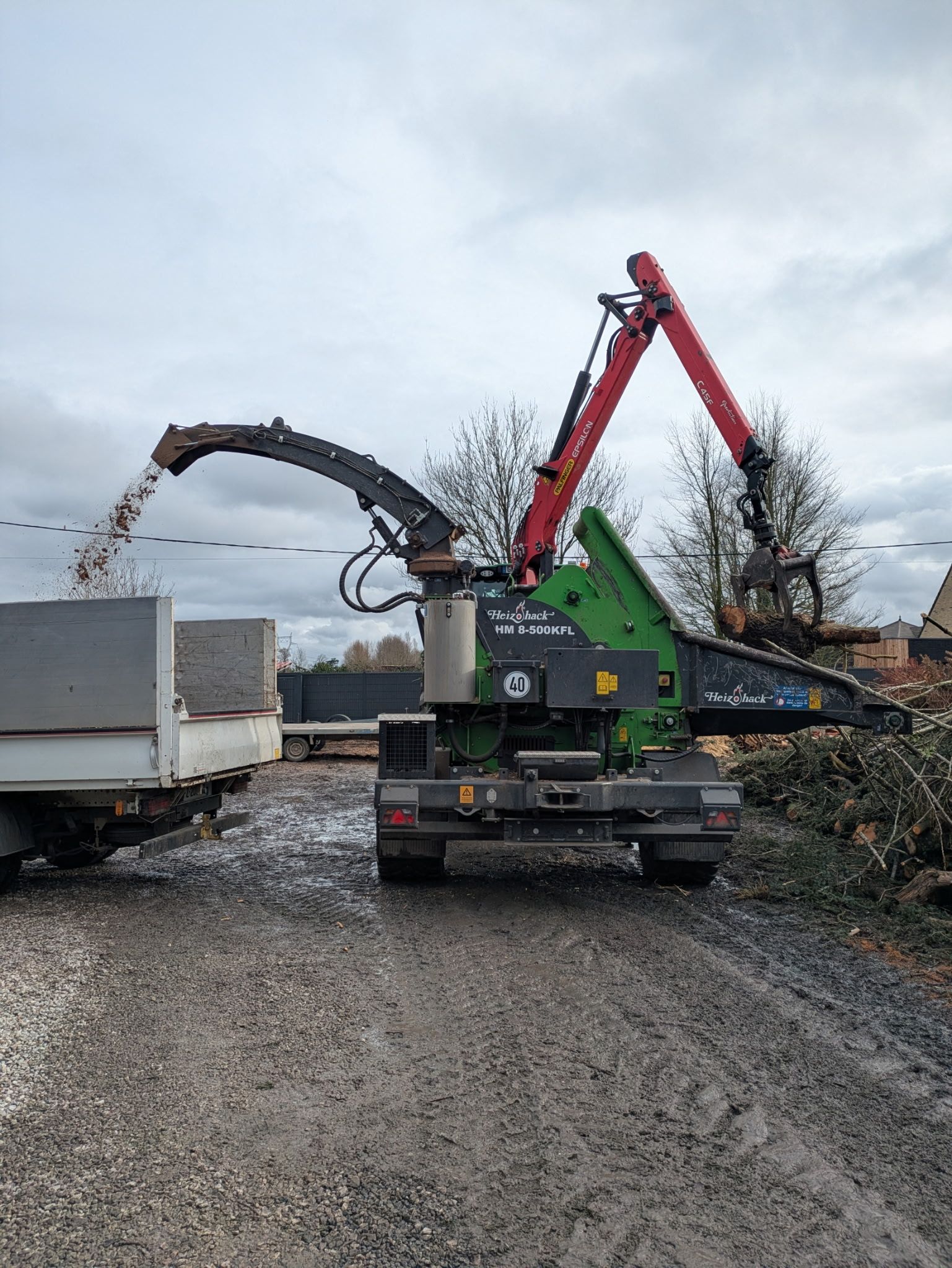 Une déchiqueteuse à bois projette des débris dans un camion blanc sur une gravière, avec un bras de flèche rouge au-dessus.