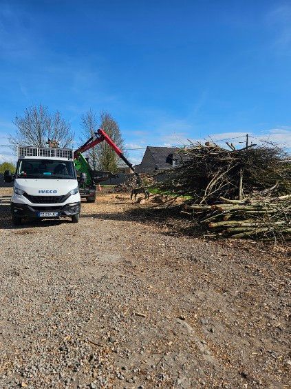 Un camion blanc et une pelleteuse rouge à côté d'un gros tas de branches d'arbres sur un terrain gravillonné