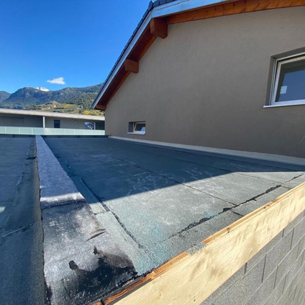 Toit plat avec membrane grise, mur d'immeuble beige et montagnes en arrière-plan.