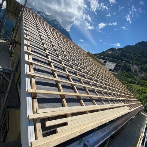 Charpente de toit en bois en construction sur un flanc de colline escarpé, avec ciel bleu et montagnes en arrière-plan.