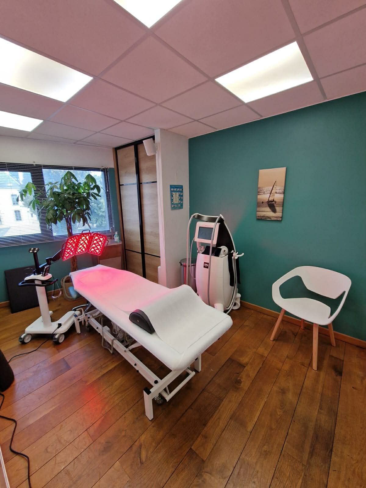Salle d'examen avec table de soins, matériel médical, mur turquoise et parquet près d'une fenêtre.